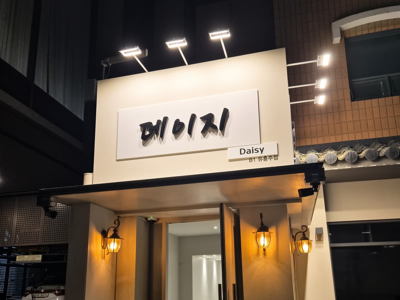 カンナム・デイジー (강남 데이지) · デイジーの看板の下に、明るい外壁と入口の両側の壁付け照明が見えます。